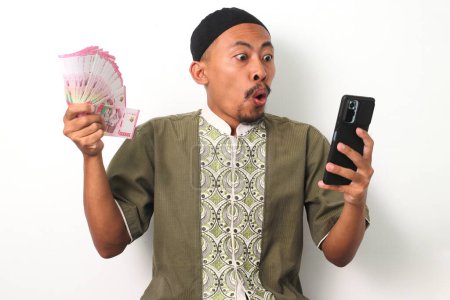 Aufgeregter indonesischer Muslim in Kokohemd und Peki hält ein Telefon und indonesische Rupiah-Banknoten in der Hand und feiert den finanziellen Erfolg. Isoliert auf weißem Hintergrund