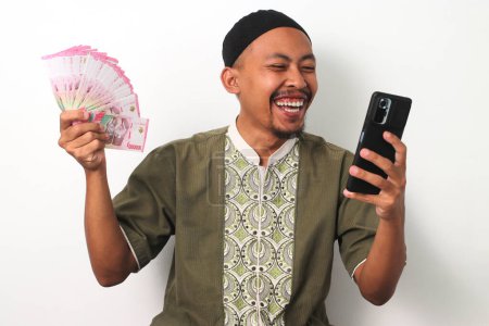 L'homme musulman indonésien excité en chemise koko et peci tient un téléphone et des billets de banque en roupie indonésienne, célébrant le succès financier. Isolé sur fond blanc