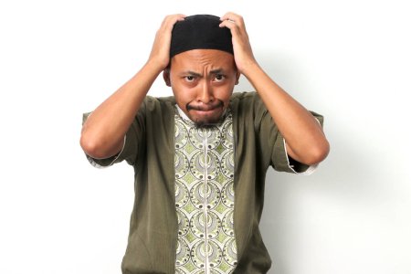 Anxious Indonesian Muslim man in koko shirt and peci grabs his head in a gesture of panic and worry. He looks at the camera with a worried expression, conveying a sense of failure. White background