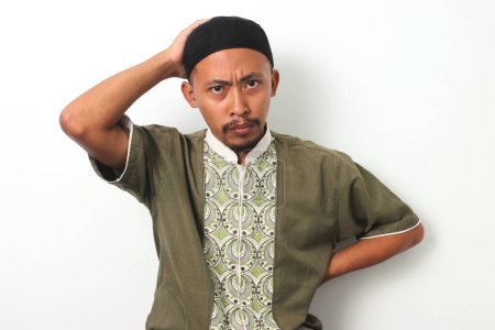 Homme musulman indonésien anxieux en chemise koko et peci attrape sa tête dans un geste de panique et d'inquiétude. Il regarde la caméra avec une expression inquiétante, véhiculant un sentiment d'échec. Fond blanc