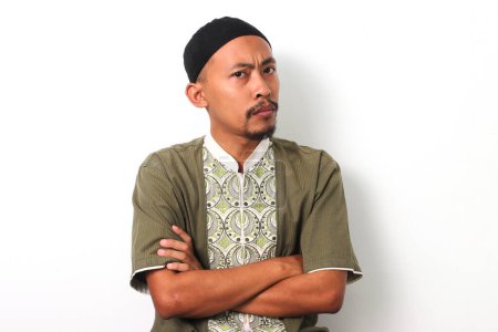 Foto de Pensativo musulmán indonesio con camisa koko y peci cruza los brazos, mirando a la cámara con una expresión dudosa o confusa. Aislado sobre fondo blanco - Imagen libre de derechos
