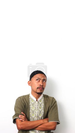 Foto de Pensativo hombre musulmán indonesio con camisa koko y peci está de pie con los brazos cruzados, reflexionando sobre una decisión. Aislado sobre fondo blanco - Imagen libre de derechos