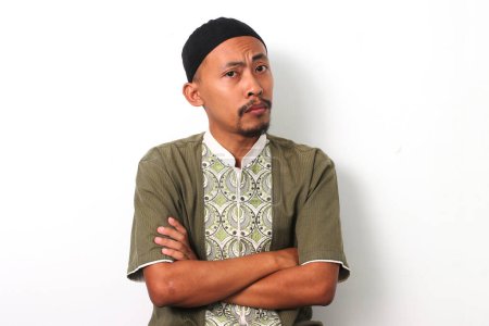 Foto de Pensativo musulmán indonesio con camisa koko y peci cruza los brazos, mirando a la cámara con una expresión dudosa o confusa. Aislado sobre fondo blanco - Imagen libre de derechos