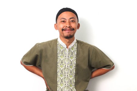 Optimistic Indonesian Muslim man in koko shirt and peci stands confidently with arms akimbo, smiling warmly at the camera. Isolated on White background