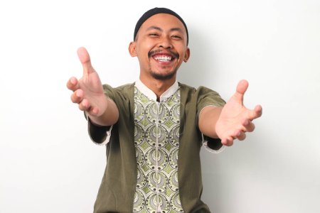 Amical, souriant homme musulman indonésien en chemise koko et peci étend ses bras pour un câlin vers la caméra, invitant une étreinte. Isolé sur fond blanc
