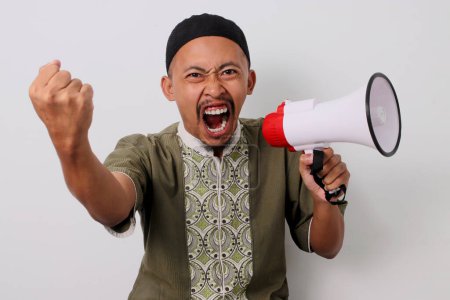 Téléchargez les photos : Homme musulman indonésien en colère à Koko et peci crie en signe de protestation, levant son poing au défi tout en tenant un mégaphone. Isolé sur fond blanc - en image libre de droit