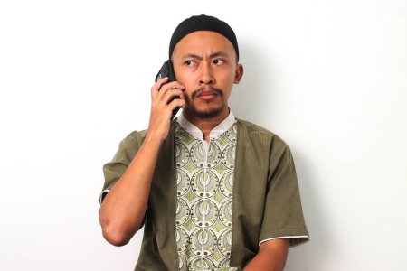 Téléchargez les photos : Homme musulman indonésien agacé à Koko et peci parle sur son téléphone, apparaissant frustré ou mécontent pendant le Ramadan. Isolé sur fond blanc - en image libre de droit