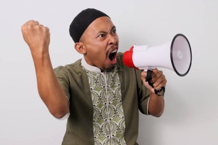 Téléchargez les photos : Homme musulman indonésien en colère à Koko et peci crie en signe de protestation, levant son poing au défi tout en tenant un mégaphone. Isolé sur fond blanc - en image libre de droit