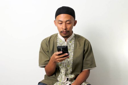L'homme musulman indonésien ennuyé à Koko et peci fait défiler son téléphone, cherchant la distraction pendant le jeûne pendant le Ramadan, en attendant l'Iftar. Isolé sur fond blanc