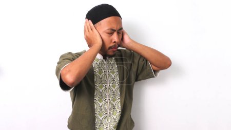 Hombre musulmán indonesio en koko y peci levanta las manos a los oídos en el gesto tradicional de realizar el Adhan (llamada a la oración). Aislado sobre un fondo blanco