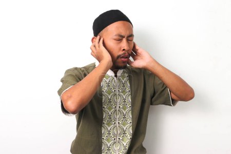 Hombre musulmán indonesio en koko y peci levanta las manos a los oídos en el gesto tradicional de realizar el Adhan (llamada a la oración). Aislado sobre un fondo blanco