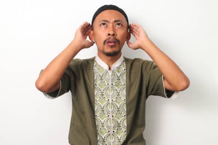 Indonesian Muslim man in koko and peci raises his hands to his ears in the traditional gesture of performing the Adhan (call to prayer). Isolated on a white background