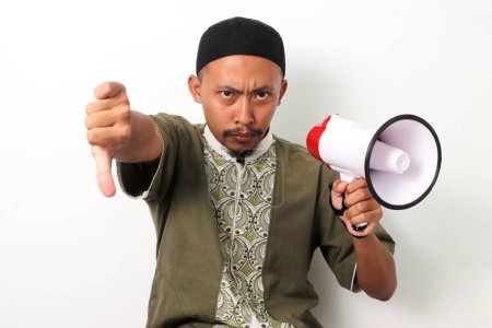 Téléchargez les photos : Homme musulman indonésien en koko et peci donne un geste de pouce vers le bas tout en tenant un mégaphone, exprimant son mécontentement ou son mécontentement. Isolé sur fond blanc - en image libre de droit