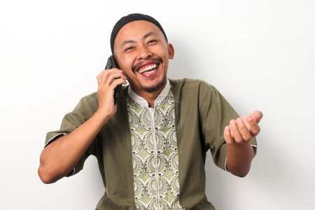 Heureux homme musulman indonésien en koko et peci parle au téléphone, riant et souriant chaleureusement lors d'une conversation agréable avec des amis ou de la famille pendant le Ramadan. Isolé sur fond blanc
