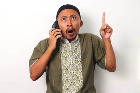 Überraschter indonesischer Muslim in Koko und Peki zeigt mit überraschtem Gesichtsausdruck auf den Kopierraum, der vor weißem Hintergrund steht