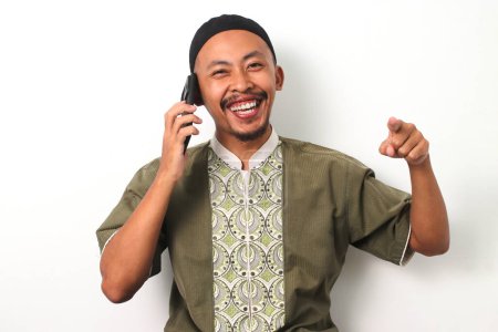 El musulmán indonesio en koko y peci señala enfáticamente la cámara mientras habla por teléfono durante el Ramadán. Aislado sobre un fondo blanco