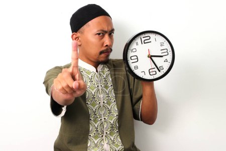 L'homme musulman indonésien en koko et peci tient une horloge et lève le doigt, signalant juste un peu plus longtemps avant que le buka puasa (brisant le jeûne du Ramadan) pendant le Ramadan. Isolé sur fond blanc