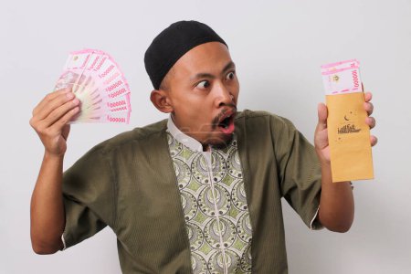 Ein überraschter indonesischer Muslim blickt auf einen braunen Umschlag voller indonesischer Rupiah-Banknoten, die sein Geldgeschenk Angpao Lebaran oder Eid al-Fitr darstellen. Isoliert auf weißem Hintergrund