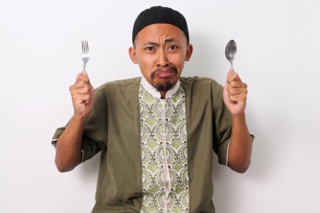 A hungry Indonesian Muslim man in koko and peci holds a fork and spoon, eagerly looking at the camera while anticipating the iftar meal to break his Ramadan fast. Isolated on a white background