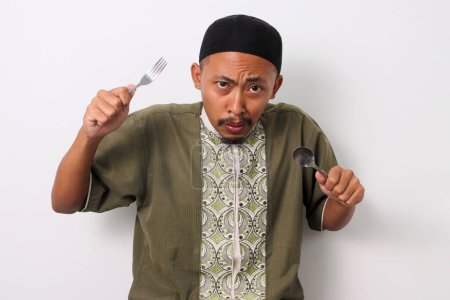 A hungry Indonesian Muslim man in koko and peci holds a fork and spoon, eagerly looking at the camera while anticipating the iftar meal to break his Ramadan fast. Isolated on a white background