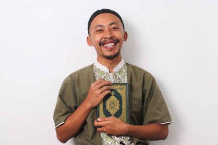 Ein fröhlicher indonesischer Muslim in Koko und Peci hält sich den Heiligen Koran an die Brust und lächelt freundlich in die Kamera. Isoliert auf weißem Hintergrund