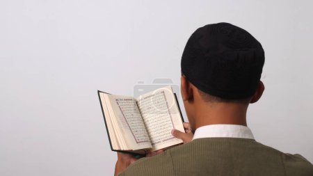 Back view of an Indonesian Muslim man in koko and peci, sitting with focus and reverence as he recites the Holy Quran during Ramadan. Isolated on a white background