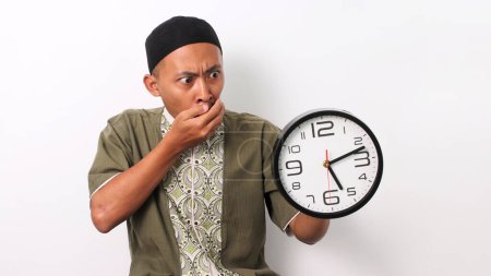 Un homme musulman indonésien choqué à Koko et peci tient une horloge, réalisant qu'il est en retard pour son repas de sahur pendant le Ramadan. Isolé sur fond blanc