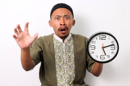 A shocked Indonesian Muslim man in koko and peci holds a clock, realizing he is late for his sahur meal during Ramadan. Isolated on a white background