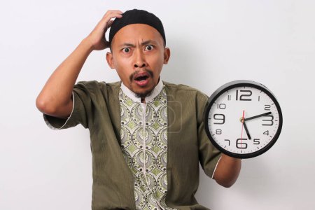 A shocked Indonesian Muslim man in koko and peci holds his head in disbelief, having realized he's late for his sahur meal during Ramadan. He holds a clock. Isolated on a white background
