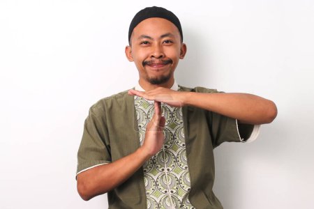 An Indonesian Muslim man in koko and peci makes the time out gesture, reminding viewers that time is running out for the sahur meal before imsak during Ramadan. Isolated on a white background