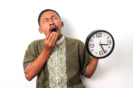 Sleepy Indonesian Muslim man in koko and peci stifles a yawn while checking a clock. He has just woken up to prepare for the suhoor meal before the Ramadan fast begins. Isolated on a white background