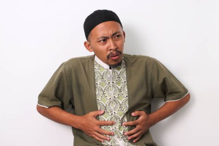 An Indonesian Muslim man in koko and peci holds his stomach with a pained expression, possibly experiencing discomfort during Ramadan. Isolated on a white background