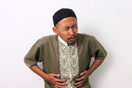 Ein indonesischer muslimischer Mann in Koko und Peki hält sich mit schmerzverzerrtem Gesichtsausdruck den Bauch und empfindet möglicherweise während des Ramadan Unbehagen. Isoliert auf weißem Hintergrund