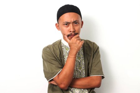 Un cher musulman indonésien en koko et peci pose sa main sur son menton, regardant attentivement la caméra pendant le Ramadan. Isolé sur fond blanc
