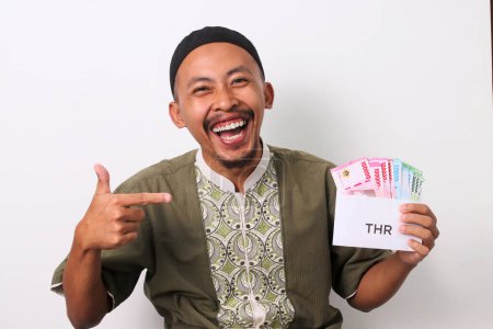 Excited Indonesian Muslim man points his finger at a white envelope labeled THR, filled with Indonesian Rupiah banknotes, representing his Religious Holiday Allowance. Isolated on a white background