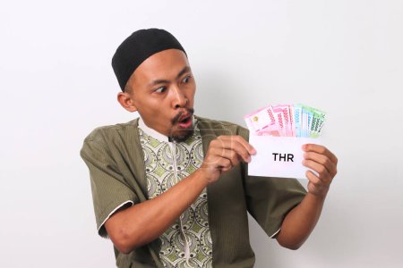 Un homme musulman indonésien choqué avec une expression surprise, tient une enveloppe blanche étiquetée THR rempli d'argent, représentant son allocation de vacances religieuses. Isolé sur fond blanc