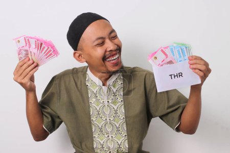 Ein aufgeregter indonesischer muslimischer Mann in Koko und Peki hält einen weißen Umschlag mit der Aufschrift THR, gefüllt mit Geld, das sein religiöses Feiertagsgeld repräsentiert. Isoliert auf weißem Hintergrund