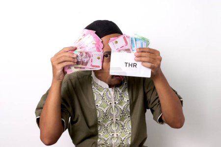 A shocked Indonesian Muslim man with a surprised expression, holds a white envelope labeled THR filled with money, representing his Religious Holiday Allowance. Isolated on a white background