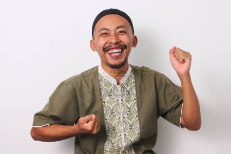 Un homme musulman indonésien joyeux à Koko et peci lève le bras et sourit pour célébrer un succès. Isolé sur fond blanc