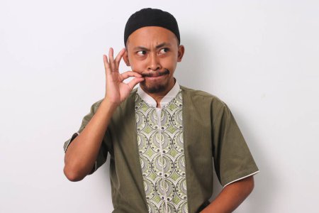 An Indonesian Muslim man makes a gesture of zipping his lips, symbolizing his commitment to mindful speech and avoiding gossip during the holy month of Ramadan. Isolated on a white background