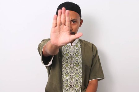 Un musulmán indonesio en koko y peci hace un gesto de alto, demostrando su compromiso de evitar prohibiciones durante el mes sagrado del Ramadán. Aislado sobre un fondo blanco