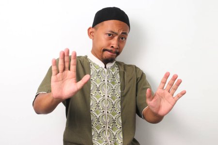 Photo for An Indonesian Muslim man in koko and peci holds up his palms in a gesture of refusal, demonstrating discipline and self-control during the Ramadan fast. Isolated on a white background - Royalty Free Image