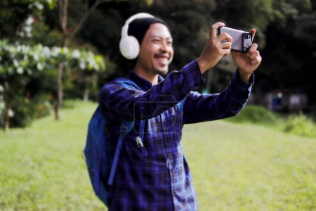 Young Asian man in a casual outfit, sporting a beanie, plaid shirt, and headphones, captures a photo on his phone during his morning nature walk. A backpack rests comfortably on his shoulder.