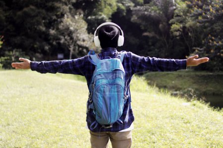 Junger asiatischer Mann in lässigem Outfit, mit Mütze, kariertem Hemd, Rucksack und Kopfhörer, breitet lächelnd die Arme aus und scheint die frische Morgenluft bei einem Spaziergang in der Natur zu genießen.