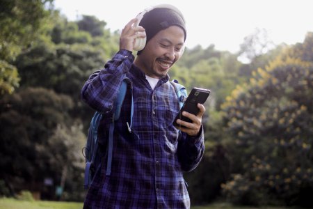 Young Asian man in a casual outfit, sporting a beanie, plaid shirt, backpack, and headphones, browses his phone for music, seemingly choosing the perfect playlist or song for his morning nature walk.
