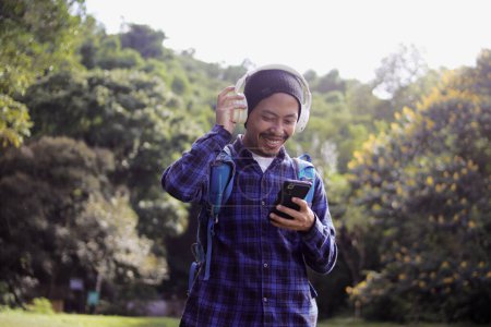 Young Asian man in a casual outfit, sporting a beanie, plaid shirt, backpack, and headphones, browses his phone for music, seemingly choosing the perfect playlist or song for his morning nature walk.