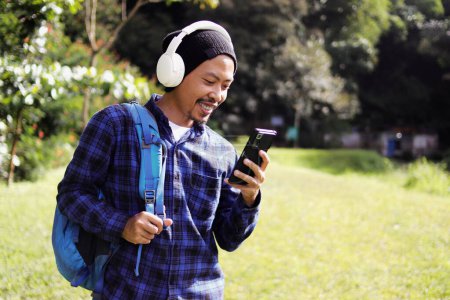 Photo for Asian man in a casual outfit, sporting a beanie, plaid shirt, and headphones, checks his phone with a grin, seemingly receiving good news while working remotely on a beautiful morning nature trail - Royalty Free Image