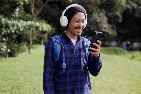 Foto de Hombre asiático en un traje casual, luciendo un gorro, camisa a cuadros y auriculares, comprueba su teléfono con una sonrisa, aparentemente recibiendo buenas noticias mientras trabaja remotamente en un hermoso sendero natural matutino - Imagen libre de derechos