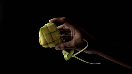 Hand presenting a ketupat pouch, a traditional Indonesian dish, woven from young coconut leaves (janur) on black background
