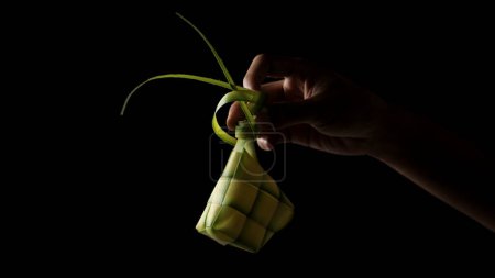 Hand presenting a ketupat pouch, a traditional Indonesian dish, woven from young coconut leaves (janur) on black background